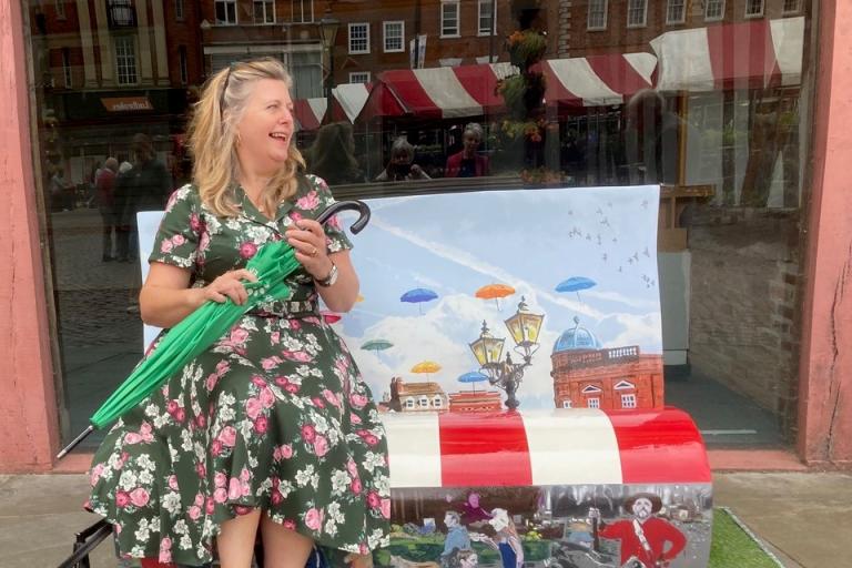 Open Doors project manager, Clare James sitting on a colorful, painted bench holding a folded umbrella