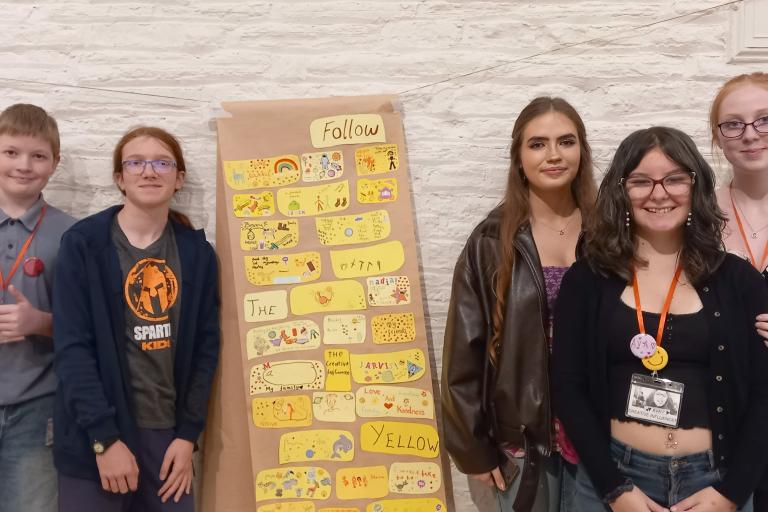 Five young people standing in a line in front of a wall decorated with a collage of drawings.