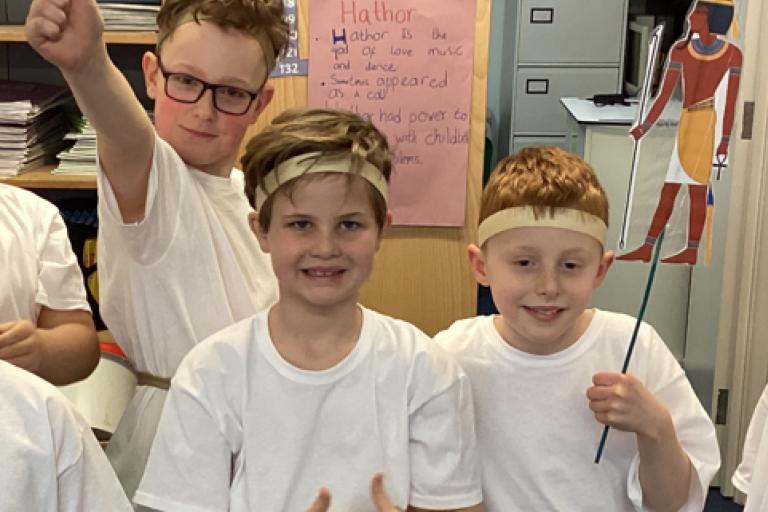 Three young boys in Ancient Egyptian costumes posing in a classroom, with one holding a cutout of an Egyptian deity.