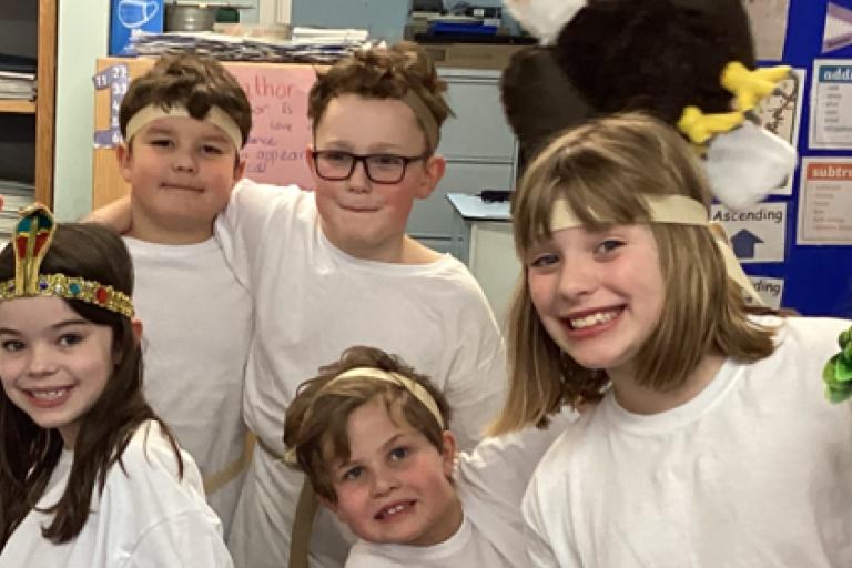 A group of children in white costumes and headbands posing for a photo in a classroom, with one wearing an eagle toy.