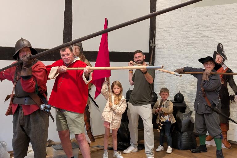 A group of adults and children posing with historical pikes, a red flag, and armour in a museum-like setting.