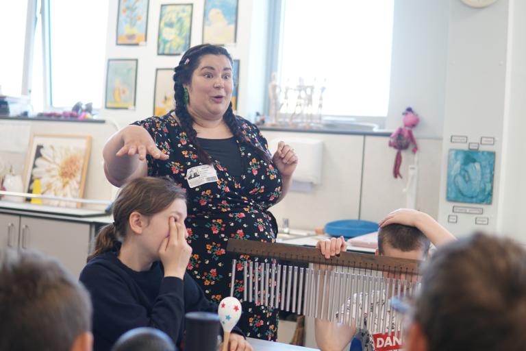 A music teacher in a floral dress gestures to students in a classroom while one student holds a bar chime instrument.