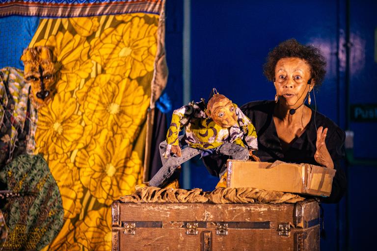 A female puppeteer in a black shirt holds up a rustic, burlap creature puppet on a dark stage.