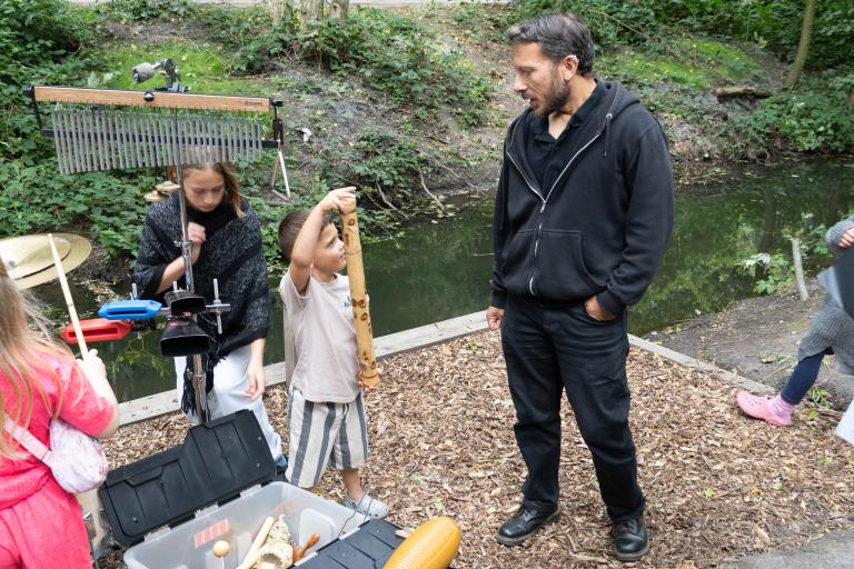A man watches a young boy playing a bamboo rainstick during an outdoor music workshop near a stream.