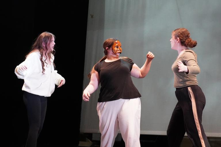 Three young women performing on a stage, with the central figure wearing a reddish-brown mask.