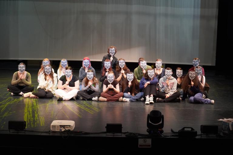 A group of young people sitting on a stage, all holding identical smiling masks in front of their faces.