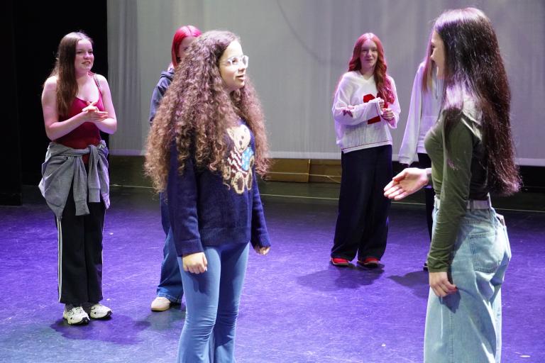A group of young teenagers rehearsing on a stage, with two girls in the foreground facing each other in conversation.
