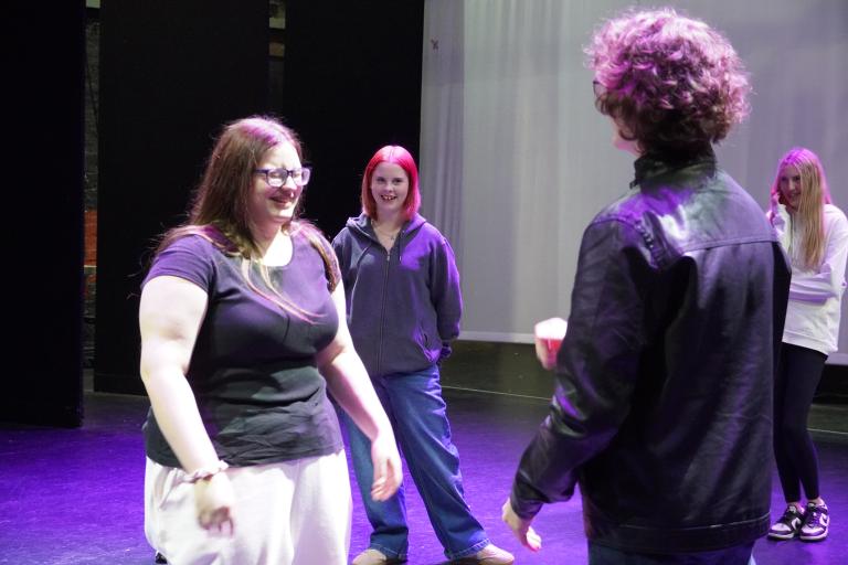 Four young people on a theatre stage under purple lighting, laughing and interacting during a rehearsal.