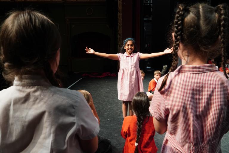A young girl in a gingham dress performs on a theatre stage with arms outstretched, viewed from behind an audience of other school children.