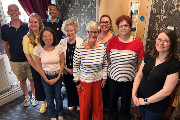 A group of nine diverse adults posing for a photo in a room with patterned wallpaper and a striped carpet.