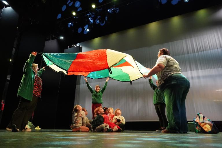 Adults holding a colourful parachute over a group of children sitting on a theatre stage.