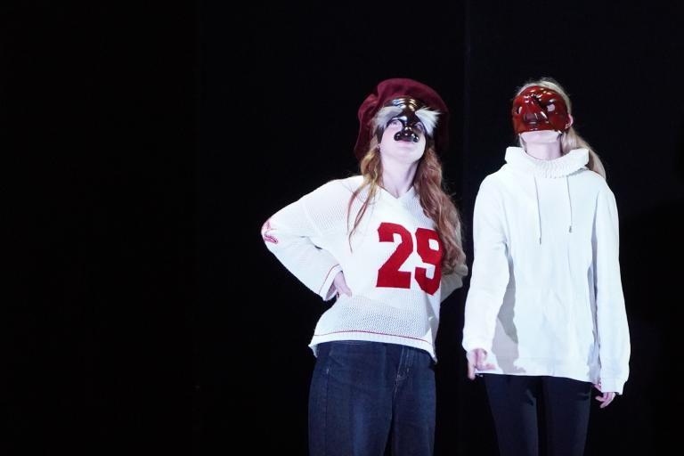 Two teenage girls in masks and casual clothing standing on a dark stage