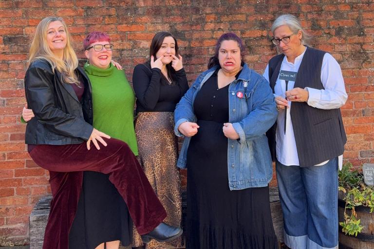 Five women posing playfully in front of a brick wall, with one holding a sign that says 'Clean me!'.
