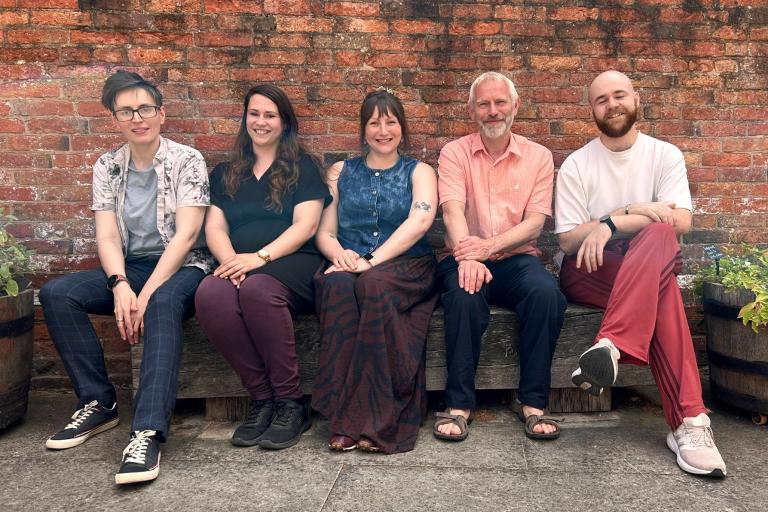 Five people sitting on a bench in front of a brick wall and smiling