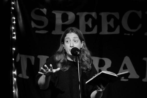 A woman performing on stage, holding an open book and gesturing while speaking into a microphone.