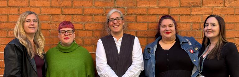 Five women standing in a row against a brick wall, dressed in varied smart-casual outfits.