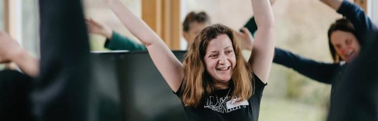 A smiling woman with brown hair and a black t-shirt participates in a group activity with her arms raised.