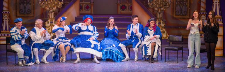 A cast of seven performers in blue and white fairytale costumes seated on stage, with two teenage presenters standing to the right.