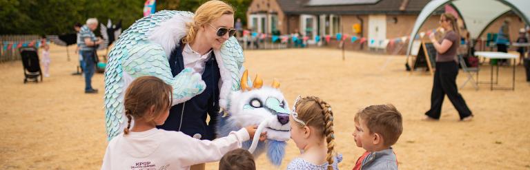 A woman in a dragon puppet costume interacts with a group of children on a grassy field at an outdoor event.