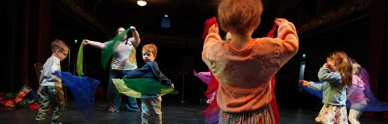 A group of children and an adult dancing with colourful fabric scarves on a dark theatre stage.