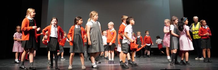 A group of primary school children in uniform performing on a theatre stage.