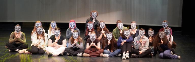 A group of young people sitting on a stage, all holding identical smiling masks in front of their faces.