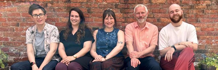 A group of five diverse people sitting in a row against a red brick wall, smiling at the camera.