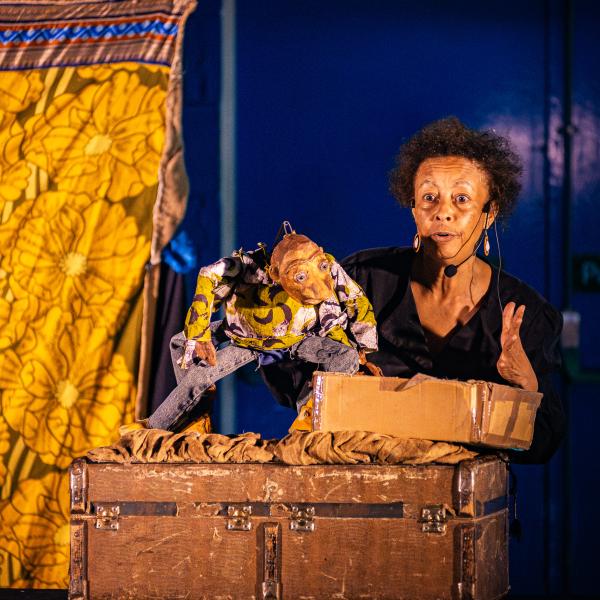 A female puppeteer in a black shirt holds up a rustic, burlap creature puppet on a dark stage.