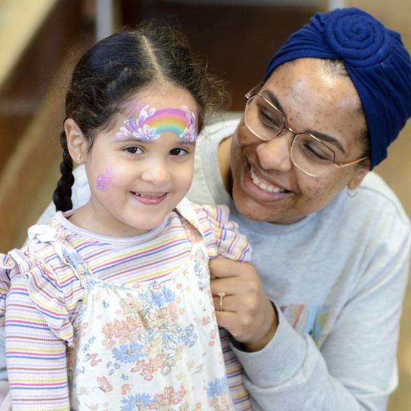 A smiling young girl with rainbow face paint on her forehead being held by a woman wearing a blue head wrap and glasses.