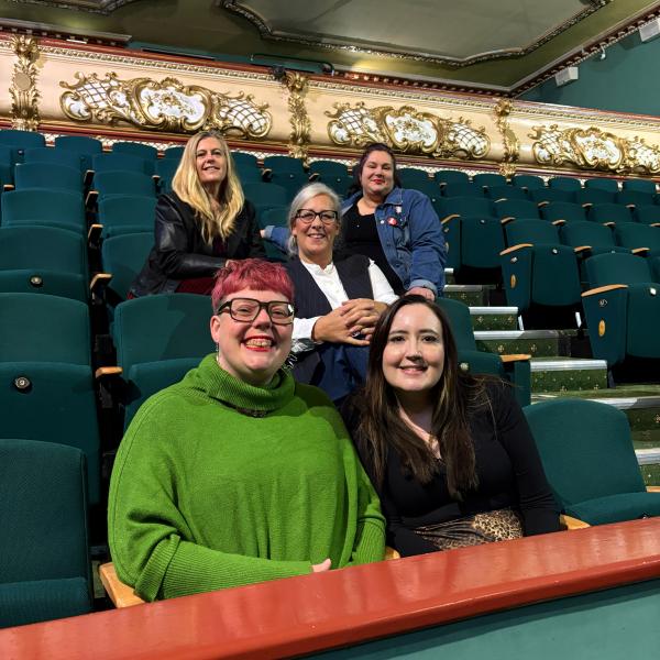 Five women posing for a photo in a theatre auditorium with dark teal seats and ornate gold balcony trim.